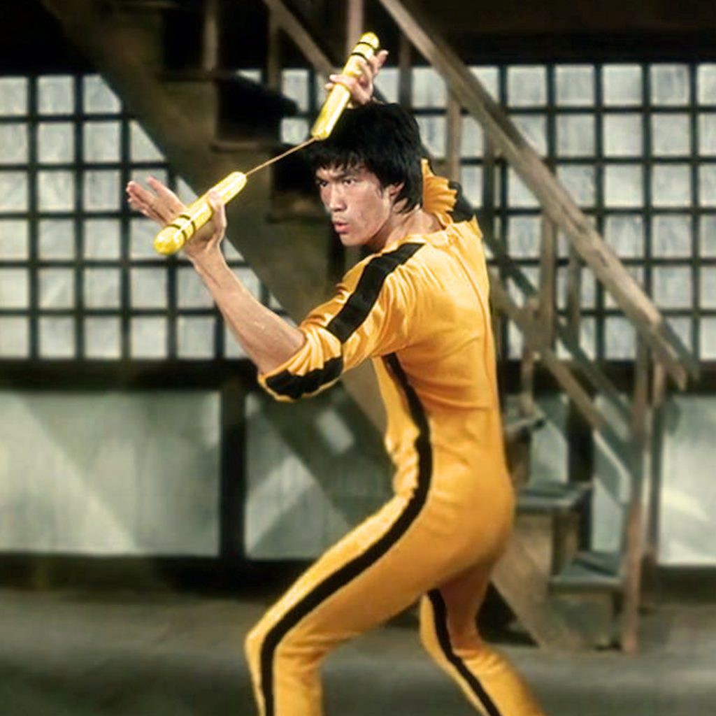 Bruce Lee in a yellow and black jumpsuit with yellow and black nunchucks